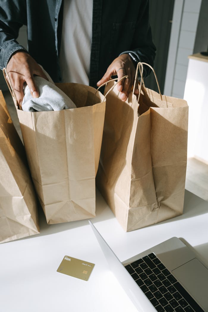 Adult organizing paper shopping bags next to a laptop and credit card on a white table.